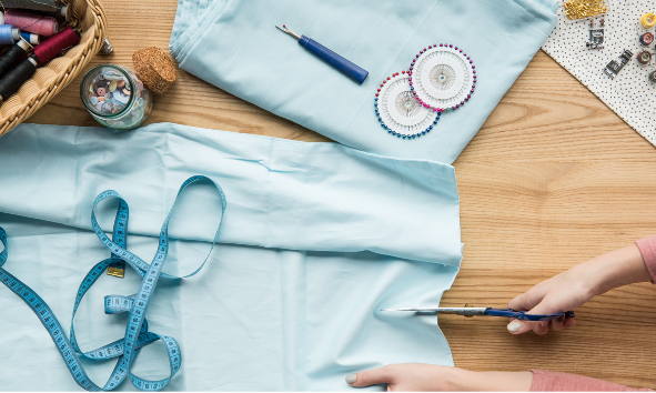 A hand cutting a cloth with scissors and measuring tape, thread, and pins surrounding it